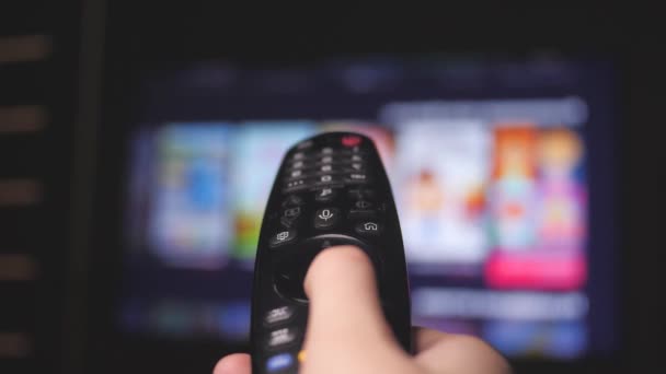 Mans mano seleziona i canali internet tv con telecomando, primo piano. La persona controlla la TV usando un moderno telecomando. Un uomo guarda la TV intelligente e usa il telecomando nero. Pagine di scorrimento tv sfocate — Video Stock