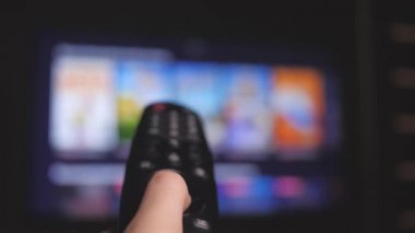 Kadın uzaktan kumandayla internet kanallarını seçiyor, yakın plan. Televizyonu modern bir uzaktan kumandayla kontrol eden kişi. Kız akıllı TV izler ve siyah uzaktan kumanda kullanır. Bulanık TV kaydırma sayfaları