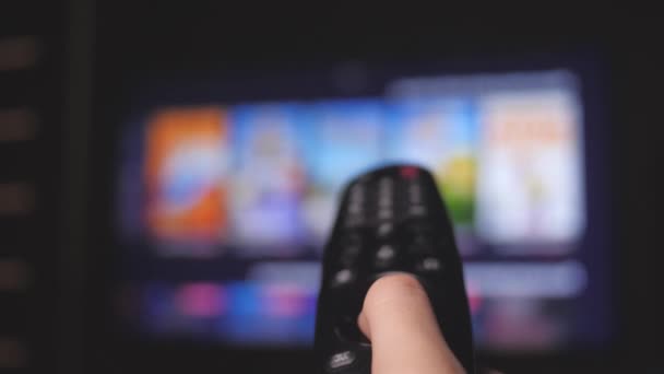 Woman hand selects internet tv channels with remote control, close-up. Person controls TV using a modern remote control. Girl watches smart TV and uses black remote control. Blurry tv scrolls pages — Stock Video