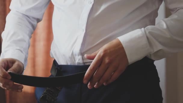 Um homem esperto põe um cinto nas calças com uma camisa branca para manter as calças nos quadris. Preparando-se de manhã cedo para o trabalho em um quarto em casa. Close-up — Vídeo de Stock