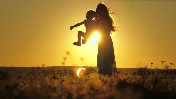 Silhouette of a mother with a small child against the sunset sky. Happy motherhood life with daughter. A woman on maternity leave walks and plays with a baby. Kid soars in the air jumping high up — Stock Photo, Image