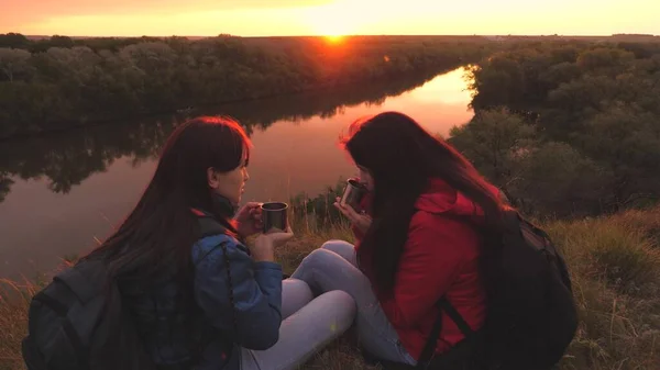 Las mujeres viajeras se sientan en lo alto de una montaña junto al río al atardecer y beben té de una taza. Ve a acampar con un amigo el fin de semana. Exploración turística del área de la naturaleza. Libérate de todos los días —  Fotos de Stock