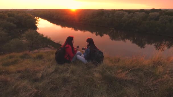 Viajantes do sexo feminino sentam-se no alto de uma montanha junto ao rio ao pôr-do-sol e bebem chá de uma caneca. Vai acampar com um amigo no fim de semana. Exploração turística da área da natureza. Livre-se de todos os dias — Vídeo de Stock