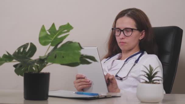 在数字电脑平板电脑上使用在线医疗应用程序微笑年轻的女性治疗师，计划在诊所办公室预约病人。快乐的医生全科医生手握触摸板 — 图库视频影像