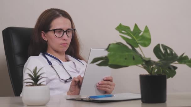Улыбающаяся молодая женщина-терапевт, использующая онлайновое приложение здравоохранения на планшете цифрового компьютера, планируя встречу с пациентами в офисе клиники. Счастливый врач общей практики держит тачпад в руках — стоковое видео