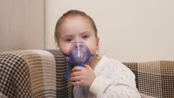 Een schattige baby met een masker ademt door een inhalator. Thuisinhalatie. Het kind krijgt ademhalingstherapie met een vernevelaar. Behandel kind van het coronavirus thuis. Een klein meisje. — Stockvideo