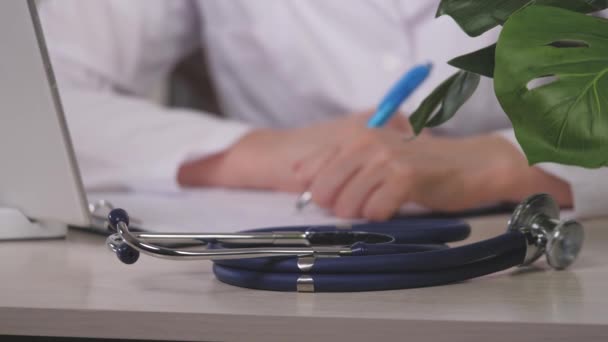 Vue rapprochée de l'équipement médical stéthoscopique avec une femme médecin travaillant en arrière-plan. Médecin professionnel gp portant des notes d'écriture manteau blanc au bureau. Concept de pratique générale — Video