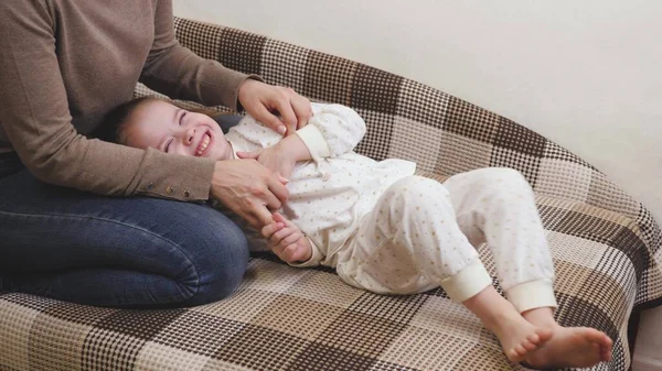 A menina brinca com sua mãe na sala das crianças no sofá e ri. Feliz vida familiar. Dia das mães. O bebê sorri enquanto está deitado ao lado da mãe. O pai faz a criança rir — Fotografia de Stock