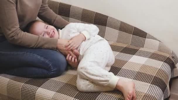 Das Mädchen spielt mit ihrer Mutter im Kinderzimmer auf dem Sofa und lacht. Glückliches Familienleben. Muttertag. Das Baby lächelt, während es neben der Mutter liegt. Die Eltern bringen das kleine Kind zum Lachen — Stockvideo