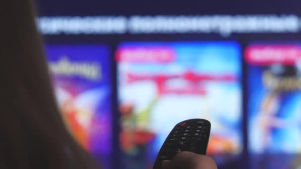 Woman hand selects internet tv channels with remote control, close-up. Person controls TV using a modern remote control. Girl watches smart TV and uses black remote control. Blurry tv scrolls pages — Stock Video