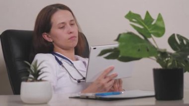 Gözlüklü yorgun kadın doktor tablet kullanarak online bir platformdan ilaç sipariş ediyor. Görme sorunu olan bir doktor kız ofiste bir koltuğa oturur ve...