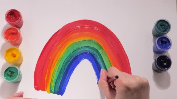 Een tiener tekent een regenboog met veelkleurige gouache verf op een wit vel papier. Kind op isolatie maakt een borsteltekening spelend met bloemen. Creativiteit in de kindertijd. Kid droomt van — Stockvideo