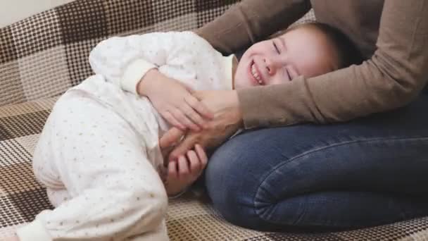 เด็กผู้หญิงเล่นกับแม่ของเธอในห้องเด็กบนโซฟาและหัวเราะ ชีวิตครอบครัวที่มีความสุข วันแม่ ทารกยิ้มขณะนอนถัดจากแม่ พ่อแม่ทําให้เด็กน้อยหัวเราะ — วีดีโอสต็อก