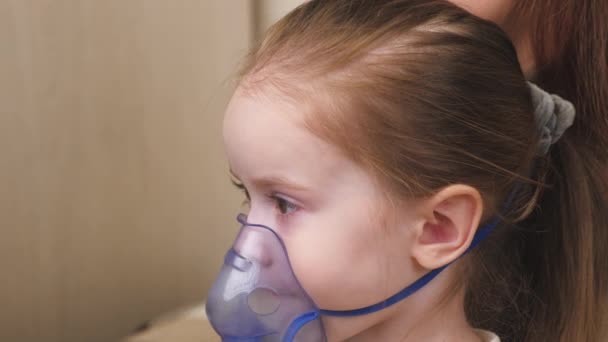 Anyu belélegez egy kislányt maszkban egy porlasztóval otthon. A gyermek légzésterápiát kap porlasztóval. Kezelje otthon a babát koronavírussal. Egy kisgyerek szenved. — Stock videók