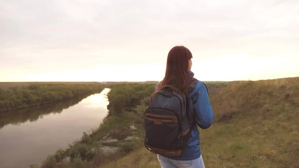 Una chica viajera con una mochila sobre sus hombros camina a lo largo del río a lo largo de una alta montaña y sonríe. Tours de senderismo. Una hermosa adolescente libre tomar un descanso del ajetreo diario y el bullicio y el estudio —  Fotos de Stock