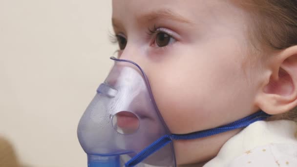Une petite fille est inhalée avec un nébuliseur dans un masque sur son visage. L'enfant reçoit un traitement respiratoire. Prévention de la grippe, des rhumes, des maladies pulmonaires et des bronchites. Gros plan d'un enfant malade. — Video
