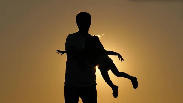 爸爸白天的轮廓与女儿在日落。女儿在父亲的怀里飞舞，胳膊伸到两边。这个女孩在她心爱的父母的怀里在空中翱翔。离职人数 — 图库照片
