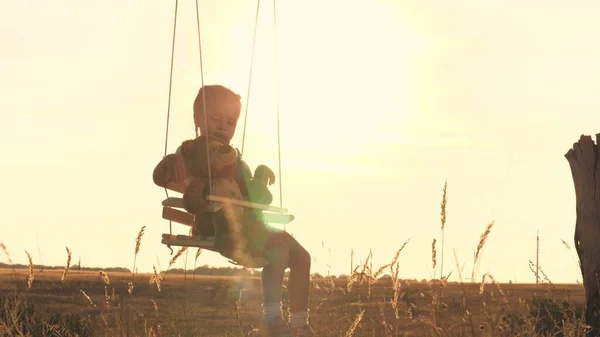 Een klein gelukkig kind rijdt hoog op een schommel met een vriend teddy in zijn armen die droomt over vliegen. Kinderen spelen buiten met een beer. De jongen zweeft tegen de hemel in de schittering van de zon. Aan — Stockfoto