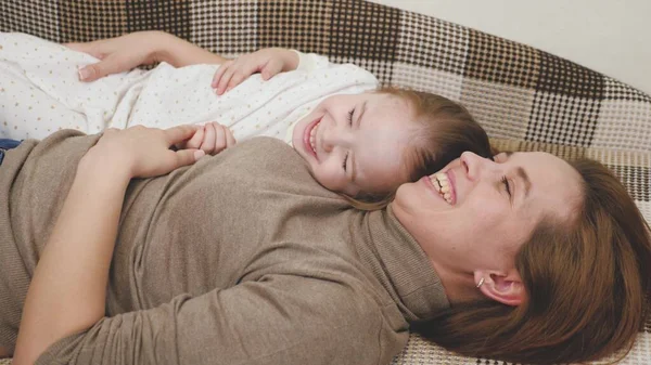 Criança brinca com a mamãe e ri enquanto está deitada no sofá. A menina feliz alegra-se com a mãe. Uma filha e uma mulher estão a brincar na sala das crianças. Dia das Mães. Os pais adoram crianças. Educação — Fotografia de Stock