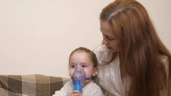 Äiti hengittää sisään pienen tytön naamiossa, jossa on sumutin kotona. Lapsi saa hengityshoitoa sumuttimella. Hoida vauvaasi sepelvaltimoviruksen varalta kotona. Pieni lapsi kärsii — kuvapankkivalokuva