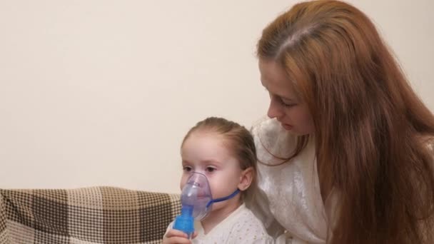 Mamma andas in en liten flicka i mask med en nebulisator hemma. Barnet får respiratorisk behandling med ett nebulisator. Behandla ditt barn för coronavirus hemma. Ett litet barn lider av — Stockvideo