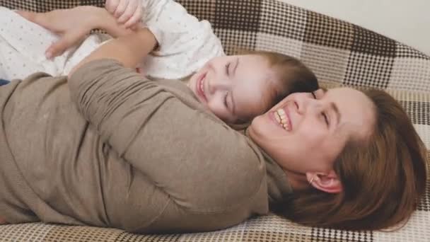 Kleines Kind spielt mit Mama und lacht, während es auf der Couch liegt. Glückliches Mädchen freut sich über Mama. Eine Tochter und eine Frau spielen im Kinderzimmer. Muttertag. Eltern lieben ihr Kind. Erziehung — Stockvideo