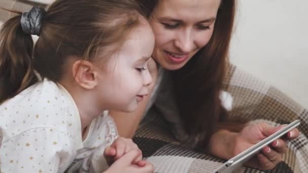 購入を選択しながら幸せなお母さんと赤ちゃんの女の子は、タブレットディスプレイを見て笑っています。ガジェットのビデオカメラを介して親戚とのリモート通信。自宅でオンラインで子供を教える — ストック動画