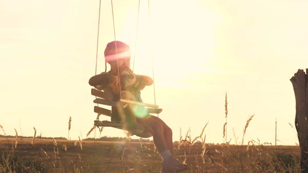 Een klein gelukkig kind rijdt hoog op een schommel met een vriend teddy in zijn armen die droomt over vliegen. Kinderen spelen buiten met een beer. De jongen zweeft tegen de hemel in de schittering van de zon. Aan — Stockfoto