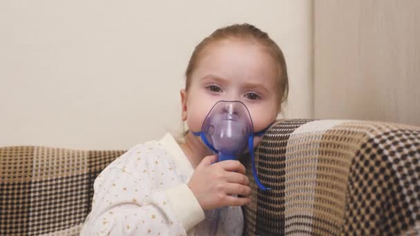 Behandling av ett barn hemma för influensa och förkylningar i lungorna. Ungen kommer att inhaleras med en nebulisator medan du sitter på soffan i barnrummet. En liten flicka andas in i en mask med medicin — Stockvideo