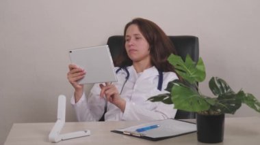 Gülümseyen genç kadın terapist dijital bilgisayar tabletinde online sağlık uygulaması kullanıyor, klinikte hasta randevusu planlıyor. Mutlu doktor pratisyen hekimin elinde dokunmatik ped var.