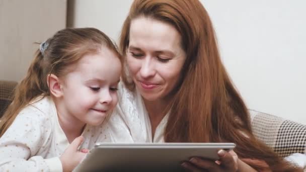 Eine glückliche Familie, Mama und Kind schauen sich einen interessanten Cartoon auf dem Tablet-Display an und lachen, das Leben spielt sich im Online-Lernen in der Anwendung ab, Mutter und Tochter liegen auf der Couch und — Stockvideo