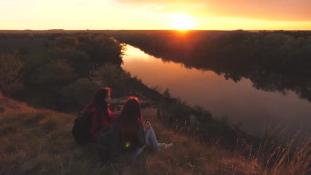 Viajantes do sexo feminino sentam-se no alto da montanha e bebem chá em uma caneca olhando para o pôr do sol. Conquistar novos lugares juntos. Fim de semana ativo. Mulheres livres turistas estão à procura de aventura na vida — Vídeo de Stock