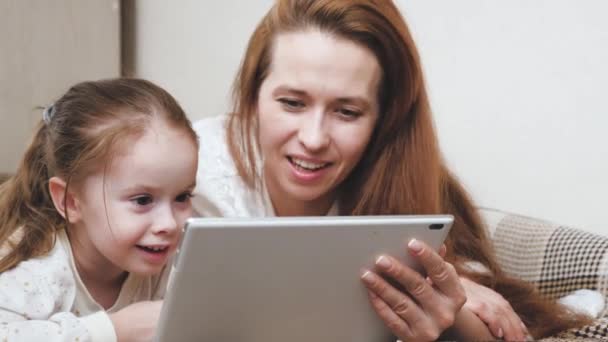 Uma mãe com uma criança pequena se comunica através da tela do tablet remotamente e ri. Mãe e filha felizes brincam juntos com um gadget enquanto estão deitados na cama. Vida familiar. Auto-isolamento em casa — Vídeo de Stock