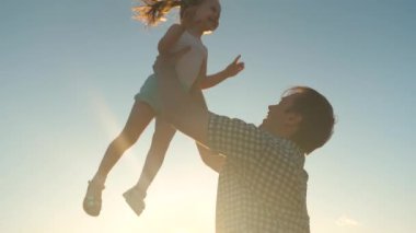 Mutlu baba güneşin parıltısıyla bir çocuğu gökyüzüne fırlatır, çocuk bir uçak gibi yükseğe uçar ve güler, küçük kız spa ile açık havada oynar, mutlu aile kavramı
