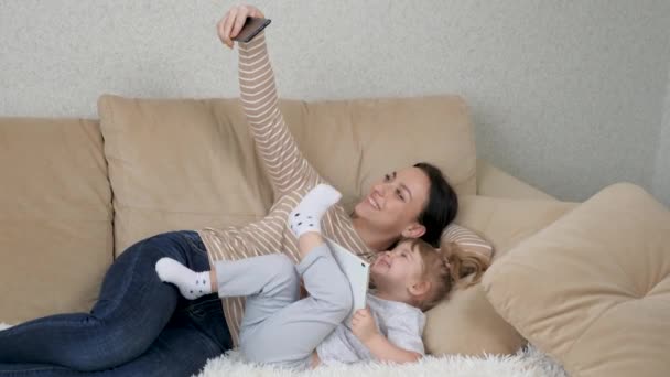 Autentyczne zbliżenie neo matki i jej dziecka wykonujących selfie lub wideo połączenie do ojca lub krewnych w łóżku. Postrzelony w 8K. Pojęcie technologii, nowe pokolenie, rodzina, więź, rodzicielstwo — Wideo stockowe