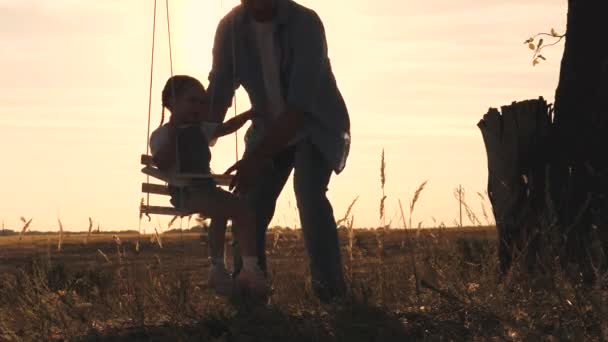 Счастливый малыш с плюшевым мишкой и папой катается на качелях на закате в парке, играя на самолёте, летящем в воздухе. Дочь и отец веселятся на рассвете. Счастливая семейная жизнь. Kid — стоковое видео