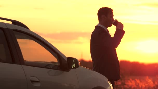 Бізнесмен у фальшивому костюмі п'є каву під час вечірнього заходу сонця в небі, що стоїть поруч з машиною, свобода подорожей за кермом, зосереджуватися і думати про майбутнє на відкритому повітрі, чоловік на світанку — стокове відео