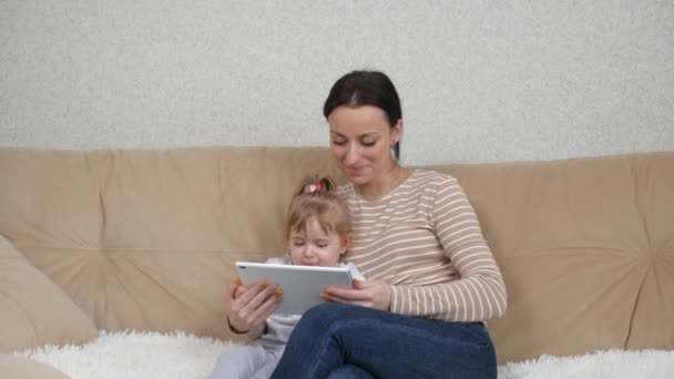 Dijital tablet kullanarak kanepede otururken gülümseyen mutlu anne ve kız çocukları. Anne ve kız kucağında ekran tableti cihazına bakıyor. — Stok video