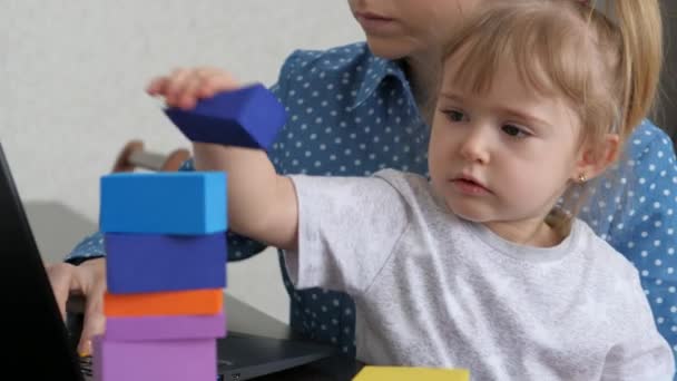 Mamma lavora in un computer portatile con un bambino piccolo tra le braccia, il bambino raccoglie una torre da cubi colorati, una donna freelance si prende cura del bambino, lavora da casa, seduto a casa, fa soldi online — Video Stock