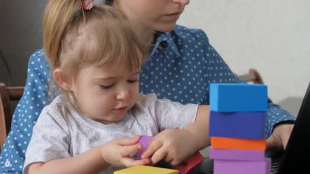 Anne kucağında küçük bir çocukla dizüstü bilgisayarda çalışıyor, çocuk renkli küplerden bir kule topluyor, bağımsız bir kadın bebeğe bakıyor, evde çalışıyor, evde oturuyor, çevrimiçi para kazanıyor. — Stok video