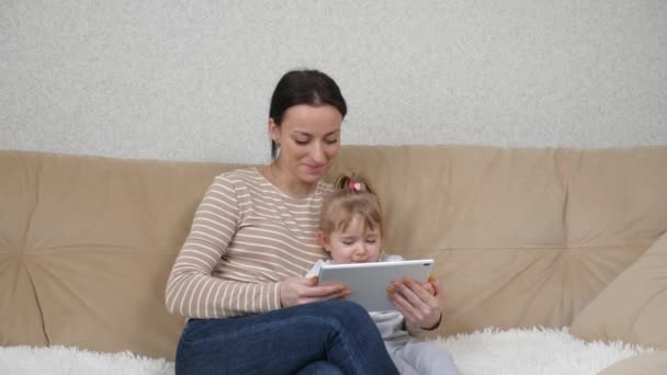 행복 한 어린 어머니와 어린 딸 이 소파에 앉아 디지털 태블릿을 사용하며 즐겁게 웃고 있습니다. 여자 아이를 안고 스크린 태블릿 장치를 보고 있는 어머니 — 비디오