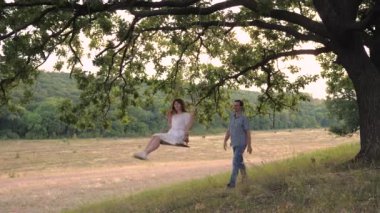 Mutlu bir kadın büyük bir meşe ağacında sallanıyor ve bir adamla gülüyor havada süzülüyor, açık havada aşk randevusu yeşil bir parkta, mutlu aile kavramı, takım çalışması, genç insanlar