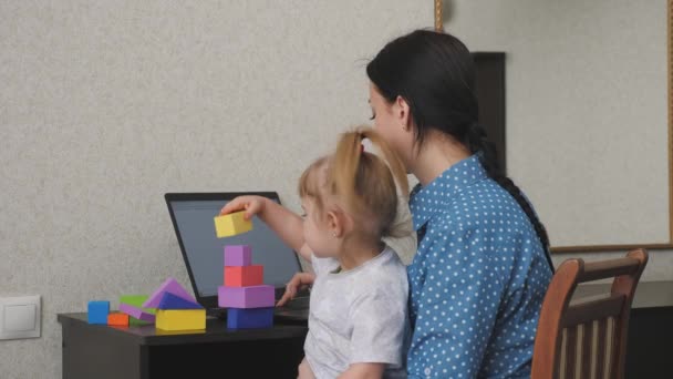 Mamma arbetar på en bärbar dator med ett litet barn i famnen, ungen samlar ett torn från färgade kuber, en frilansande kvinna tar hand om barnet, arbetar hemifrån, sitter hemma, tjäna pengar på nätet — Stockvideo