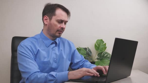 Un hombre serio trabaja en una computadora portátil en la oficina, un empleado lleva a cabo un negocio en una computadora, un freelancer escribe documentos importantes en un teclado, pagos por recibos en línea a través de Internet — Vídeo de stock