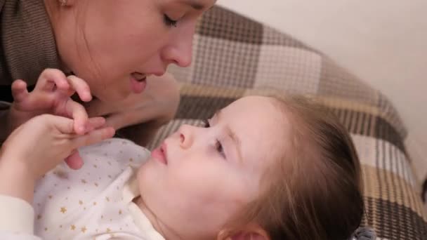 Little happy girl hugs her beloved mother and kisses, the child with the mother lie on the sofa in the childrens room and laugh, parental affection and support for the kid, the concept of family — Stock Video