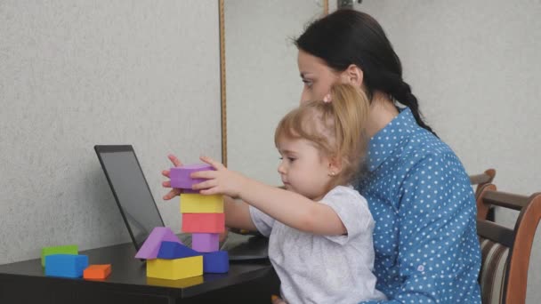 Mam werkt op een laptop met een klein kind in haar armen, het kind verzamelt een toren van gekleurde blokjes, een freelance vrouw zorgt voor de baby, werkt vanuit huis, zit thuis, geld verdienen online — Stockvideo