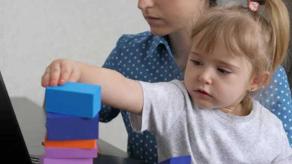 Mom works at a laptop with a small child in her arms, the kid collects a tower from colored cubes, a freelance woman takes care of the baby, working from home, sitting at home, making money online