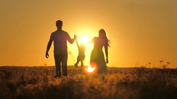 Silhouette of a private family at sunset in the sky, mother, father and child walk outdoors, girl flies in the arms of dad and mom at dawn, travel in the evening in the glare of the sun in a beautiful