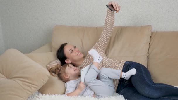 Mor med et lille barn tager en selfie på en smartphone, mens de ligger på sofaen og griner, familie fotosessioner hjemme indendørs, mor og datter, lykkelige i livet, har en god tid sammen. – Stock-video