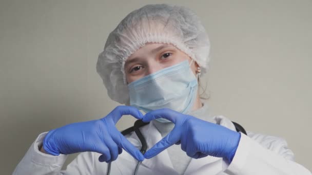 Un médecin avec un masque bleu et des gants sourit et montre son cœur, un cardiologue professionnel dans une clinique de santé, un rendez-vous avec un pédiatre au bureau, une bonne infirmière, une pandémie de coronavirus — Video